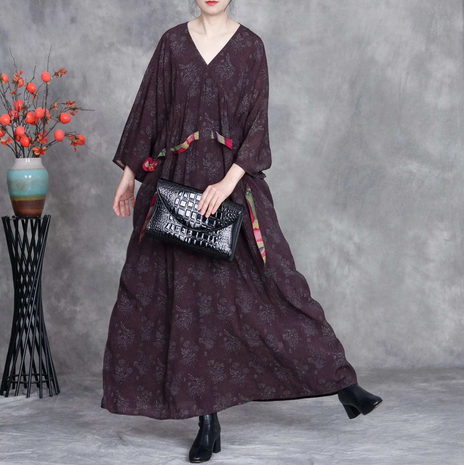 Woman wearing a long dark patterned dress holding a black handbag against a gray background.
