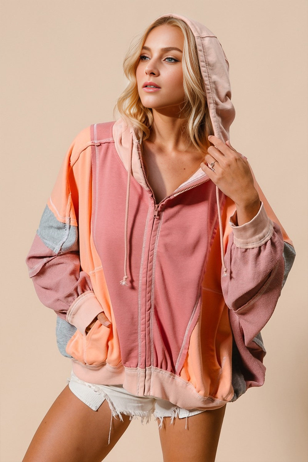 A woman wearing a pink and orange color block zip up hoodie with a drawstring hood, paired with white shorts.