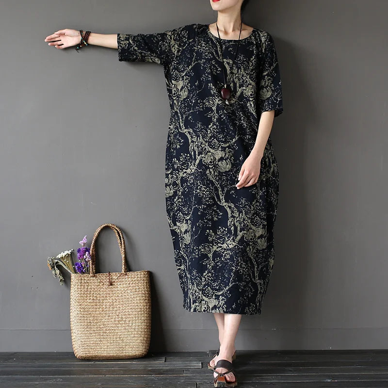 Woman wearing a dark floral dress standing next to a woven bag against a gray wall.