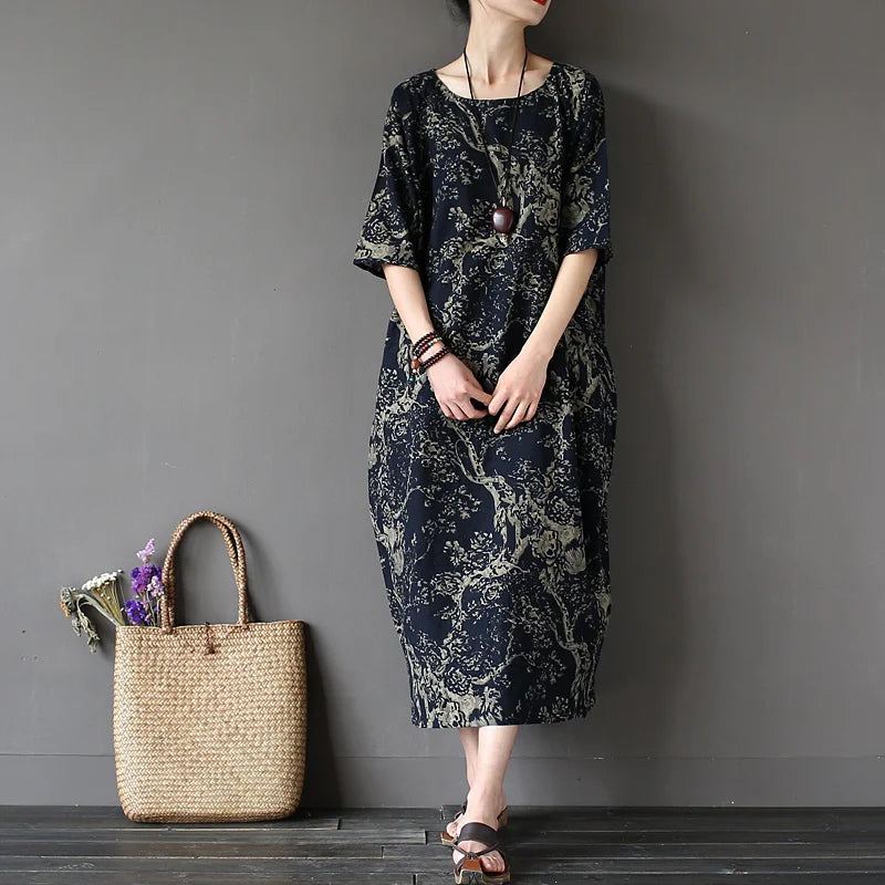 Woman wearing a dark floral dress standing next to a woven bag against a gray wall.