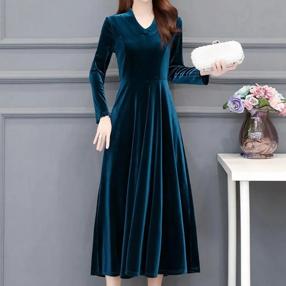 Woman wearing a teal velvet dress holding a white clutch against a gray wall.