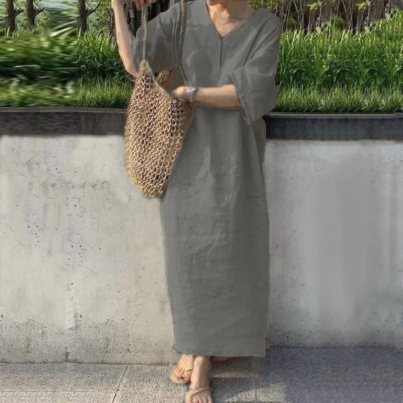 Person wearing a long gray dress holding a woven bag against a concrete wall with greenery.