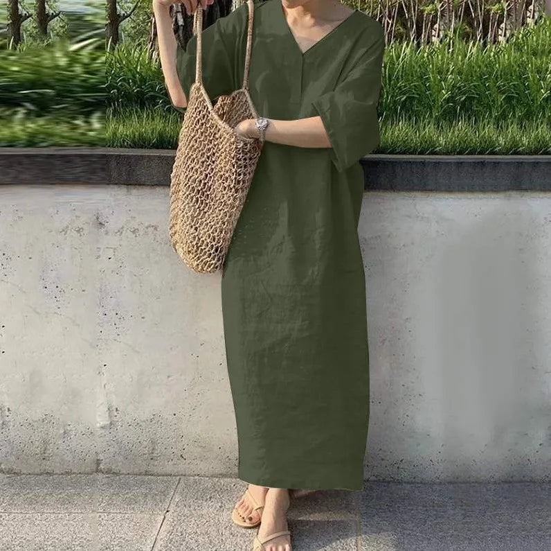 Person wearing a green dress holding a woven bag against a concrete wall with greenery.