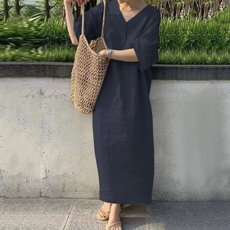 Person wearing a dark blue dress holding a woven bag against a concrete wall with greenery.