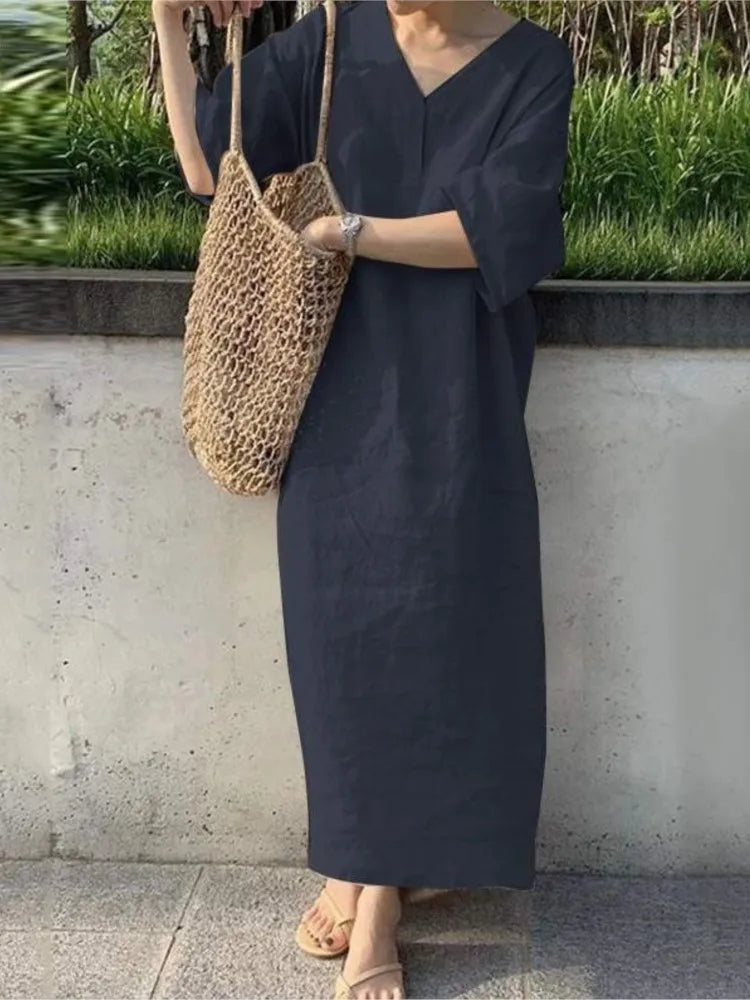 Person wearing a dark blue dress holding a woven bag against a concrete wall with greenery.