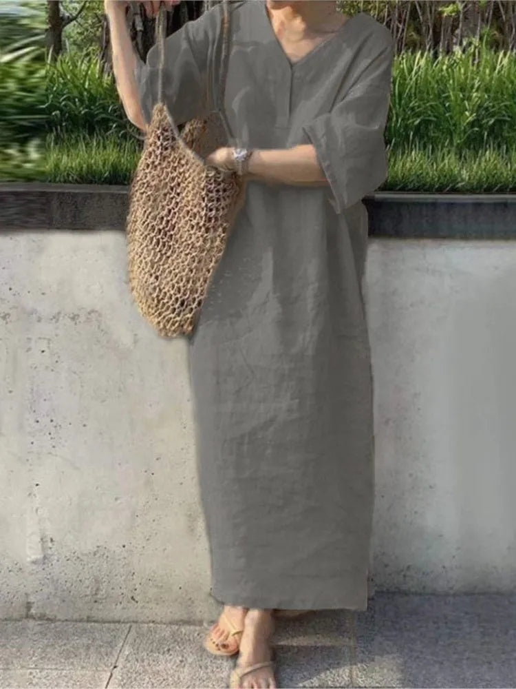 Person wearing a long gray dress holding a woven bag against a concrete wall with greenery.