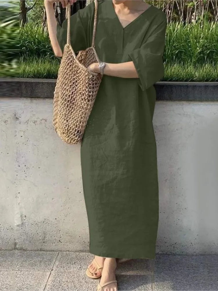 Person wearing a green dress holding a woven bag against a concrete wall with greenery.