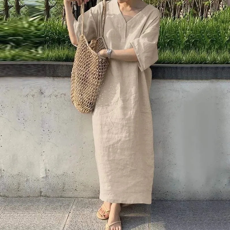 Person wearing a beige dress holding a woven bag against a concrete wall with greenery.