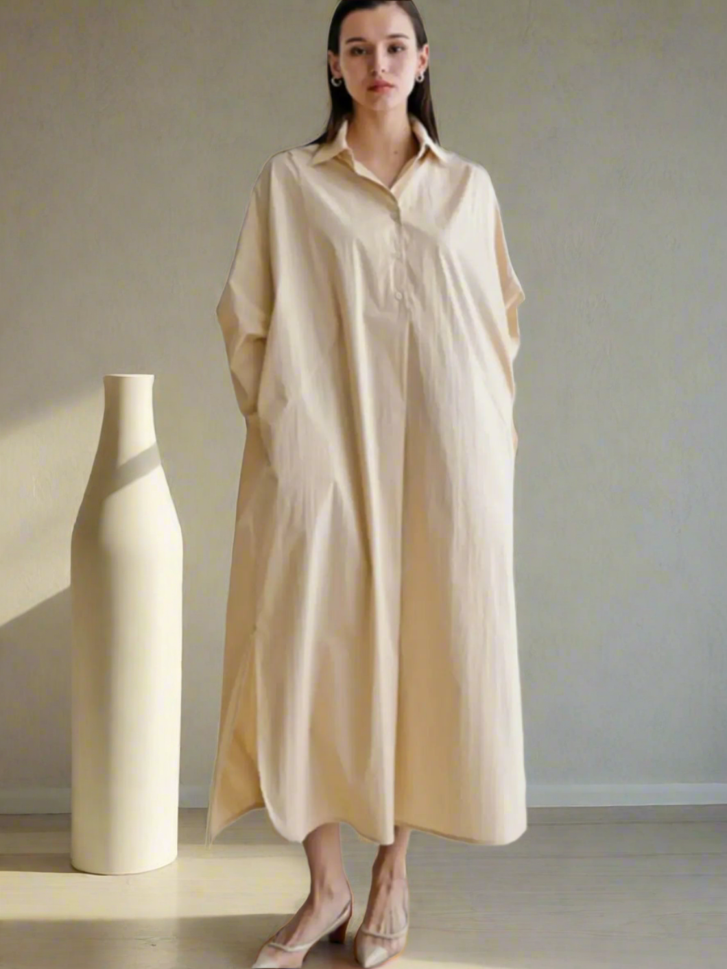Woman wearing a beige long dress standing next to a white vase against a plain background