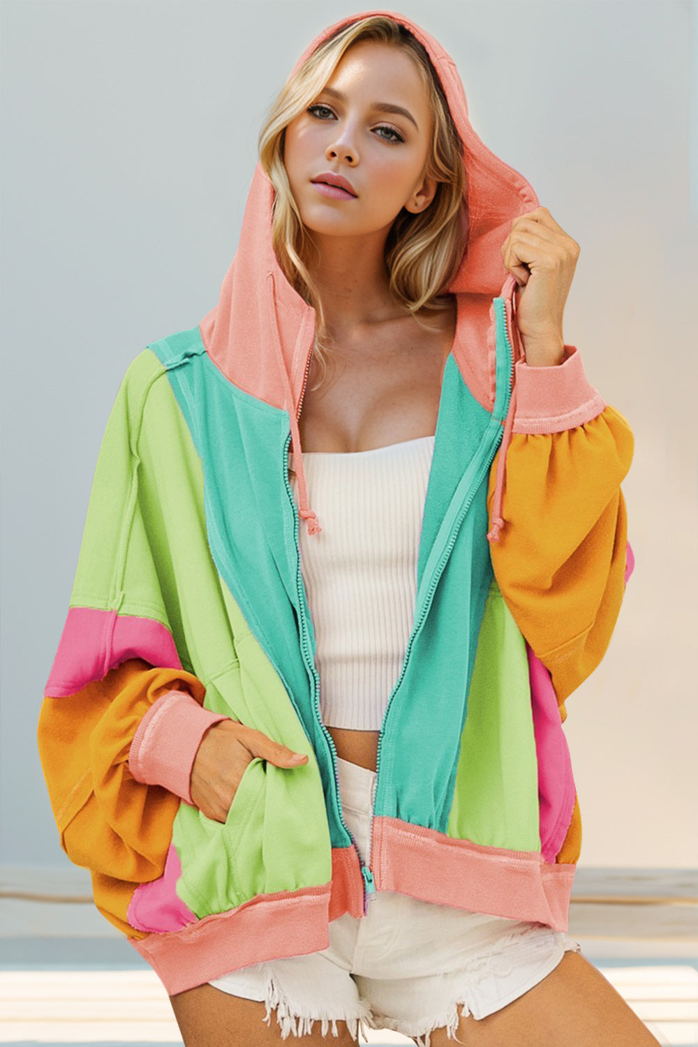 A woman wearing a color block zip up hoodie with hood, featuring pink, green, yellow, and orange colors, paired with white shorts.