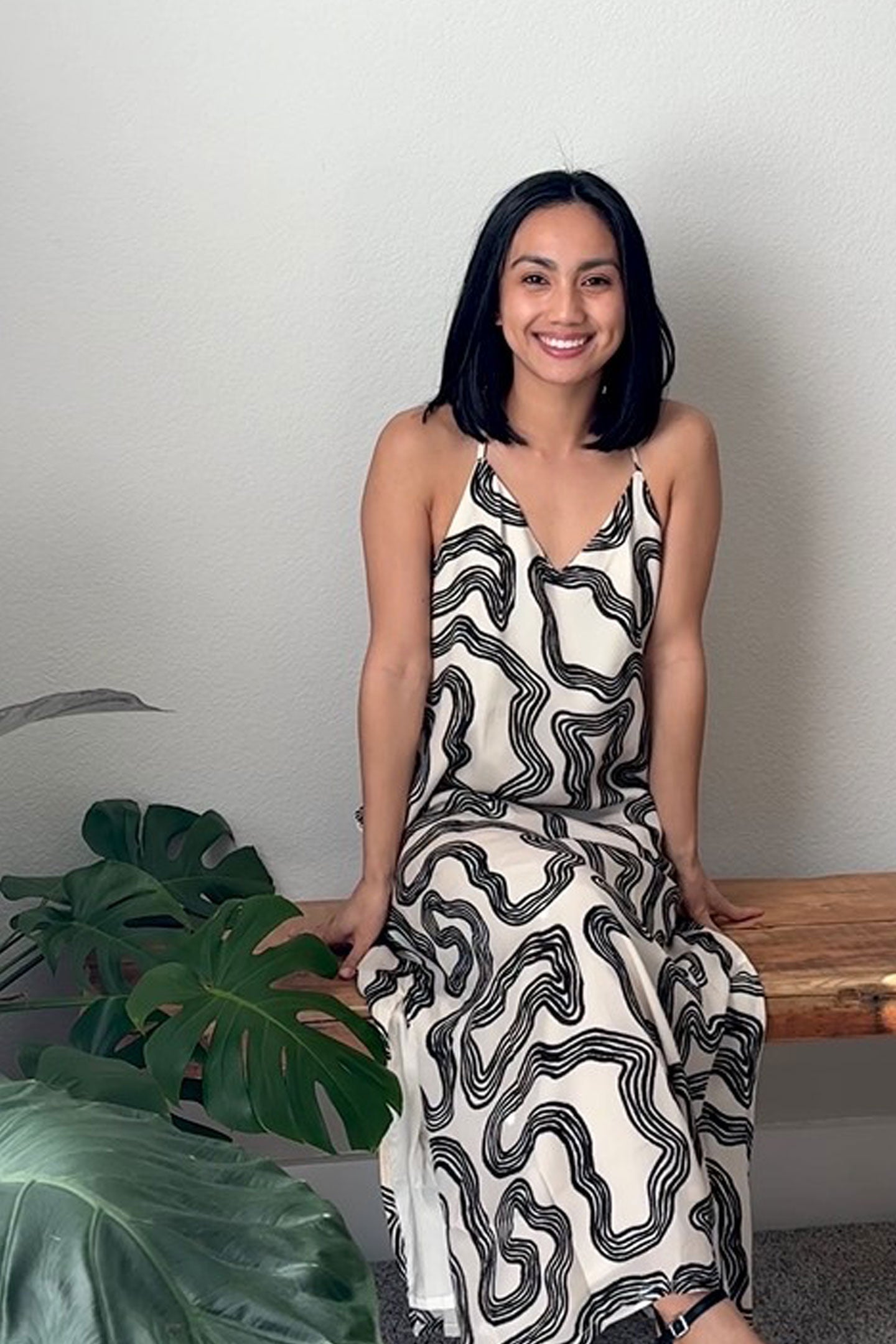 Woman wearing a black and white patterned dress sitting on a wooden bench with plants in the background.
