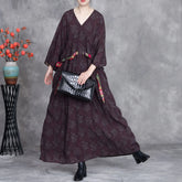 Woman wearing a long dark patterned dress holding a black handbag against a gray background.