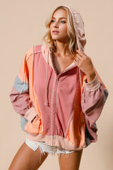 A woman wearing a pink and orange color block zip up hoodie with a drawstring hood, paired with white shorts.