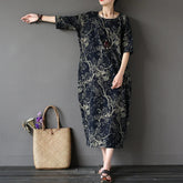 Woman wearing a dark floral dress standing next to a woven bag against a gray wall.