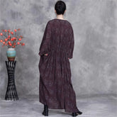 Person wearing a long dark purple dress standing in front of a gray wall with a decorative plant on a stand.