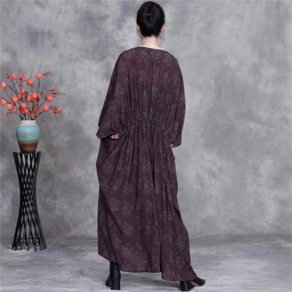 Person wearing a long dark purple dress standing in front of a gray wall with a decorative plant on a stand.