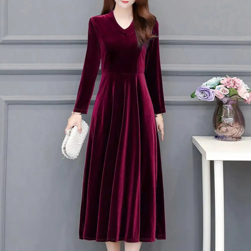 Woman wearing a burgundy velvet dress against a gray wall with a small table and flowers.