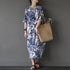 Woman wearing a blue and white floral dress holding a woven bag against a gray background