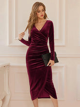 Woman wearing a burgundy velvet dress holding a black clutch against a white wall.