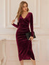 Woman wearing a burgundy velvet dress holding a black clutch against a white wall.