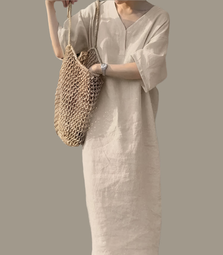 Person wearing a beige dress holding a woven bag against a concrete wall with greenery.
