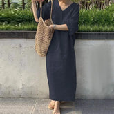 Person wearing a dark blue dress holding a woven bag against a concrete wall with greenery.