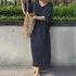 Person wearing a dark blue dress holding a woven bag against a concrete wall with greenery.