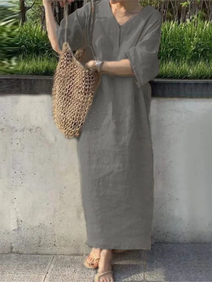 Person wearing a long gray dress holding a woven bag against a concrete wall with greenery.