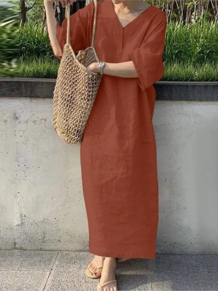 Person wearing a rust-colored dress holding a woven bag against a concrete wall with greenery.