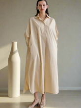 Woman wearing a beige long dress standing next to a white vase against a plain background