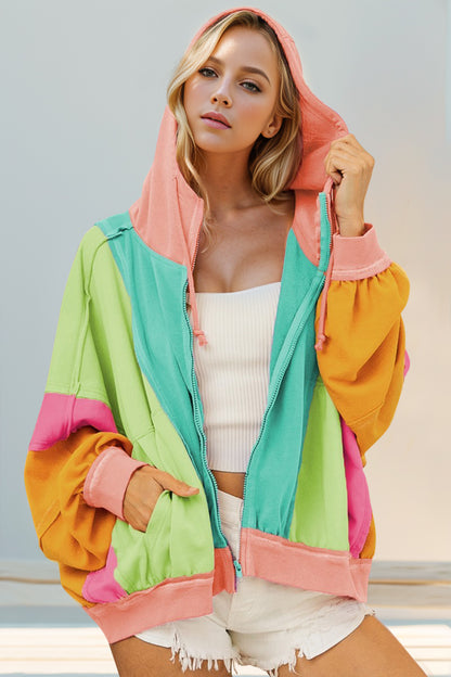 A woman wearing a color block zip up hoodie with hood, featuring pink, green, yellow, and orange colors, paired with white shorts.